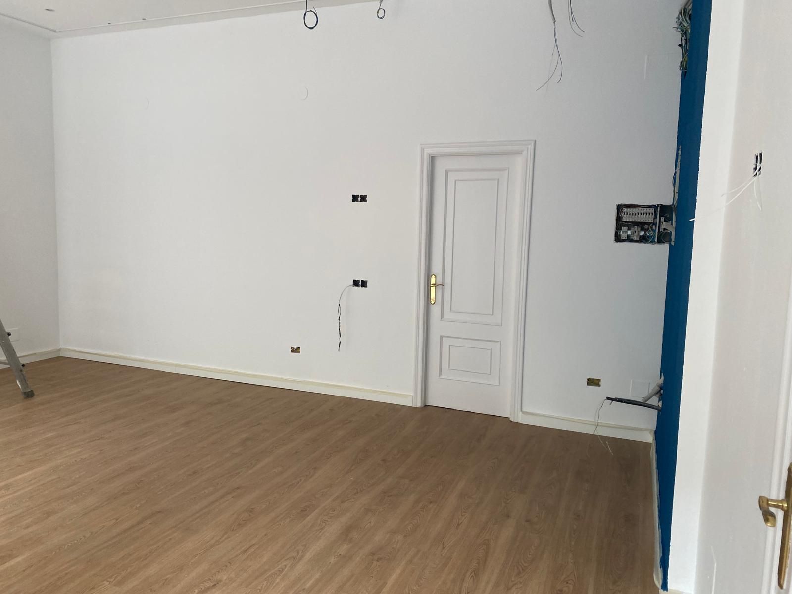 Habitación vacía con paredes blancas, suelo de madera y una pequeña puerta blanca cerca del centro.