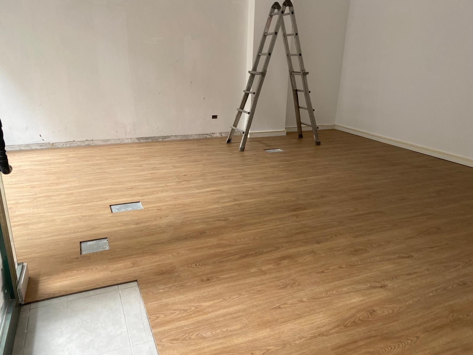 Habitación vacía con suelo de madera, una escalera apoyada contra la pared y dos enchufes en el suelo a la vista.