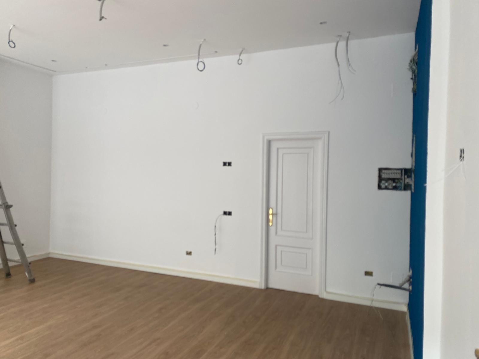Habitación blanca vacía con suelo de madera, una puerta pequeña, cables a la vista en el techo y una escalera a la izquierda.