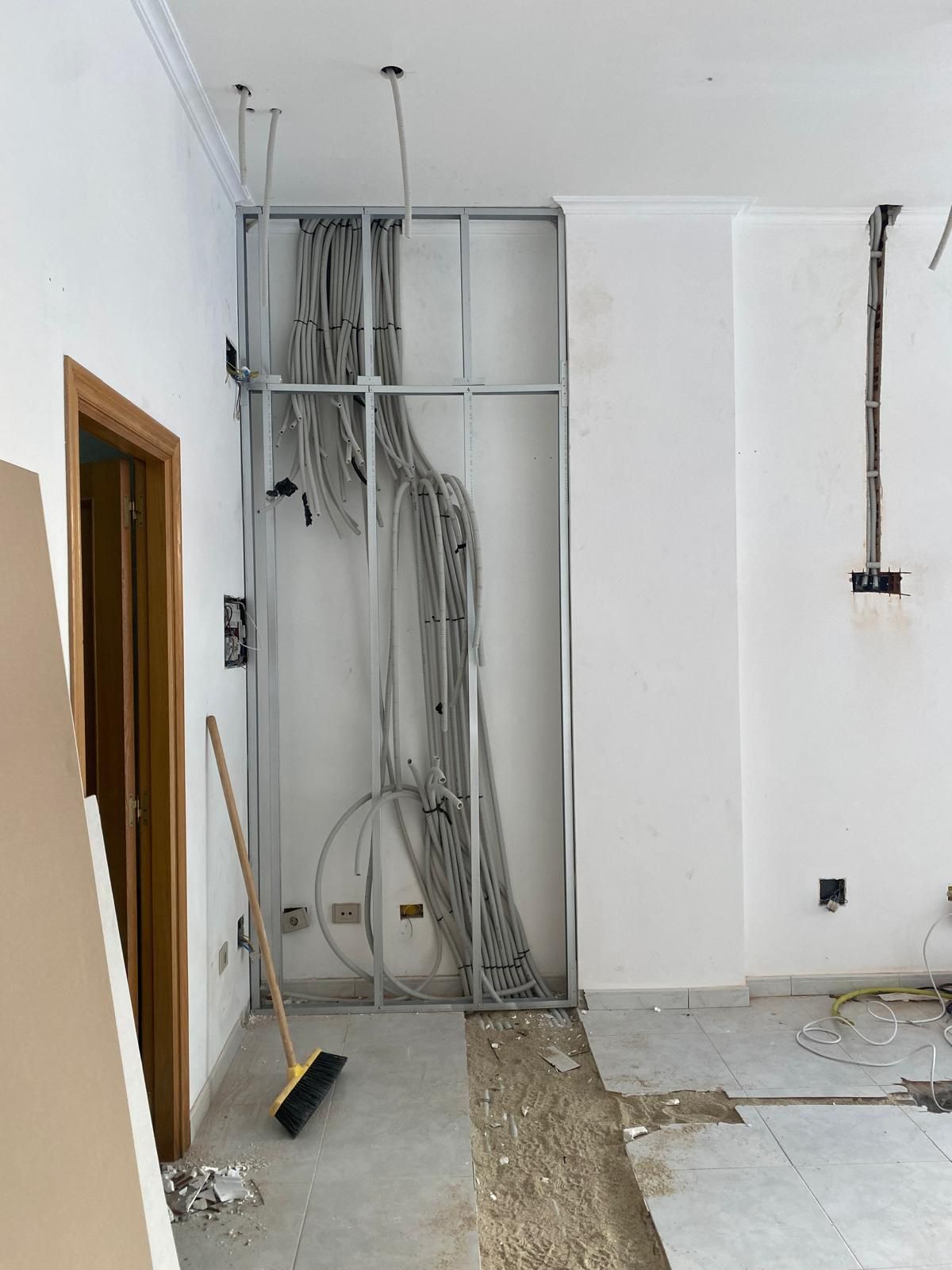 Habitación parcialmente terminada con cableado eléctrico a la vista, paredes blancas y una puerta a la izquierda.