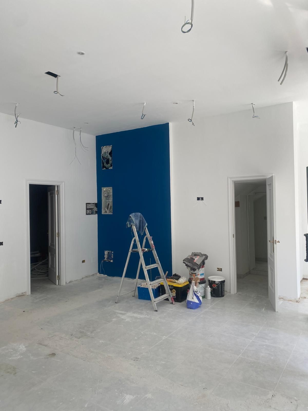 Habitación sin terminar con una pared decorativa azul, escaleras, cubos de pintura y cables del techo a la vista.