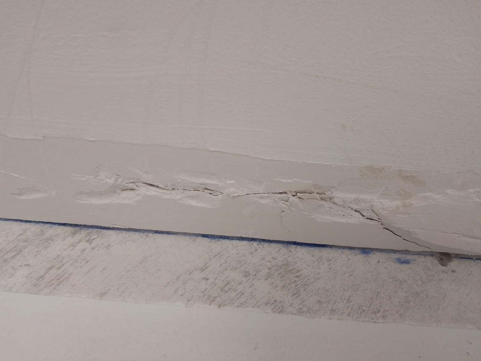 Pintura blanca descascarada y yeso por encima de una línea de cinta azul en una esquina de pared dañada.