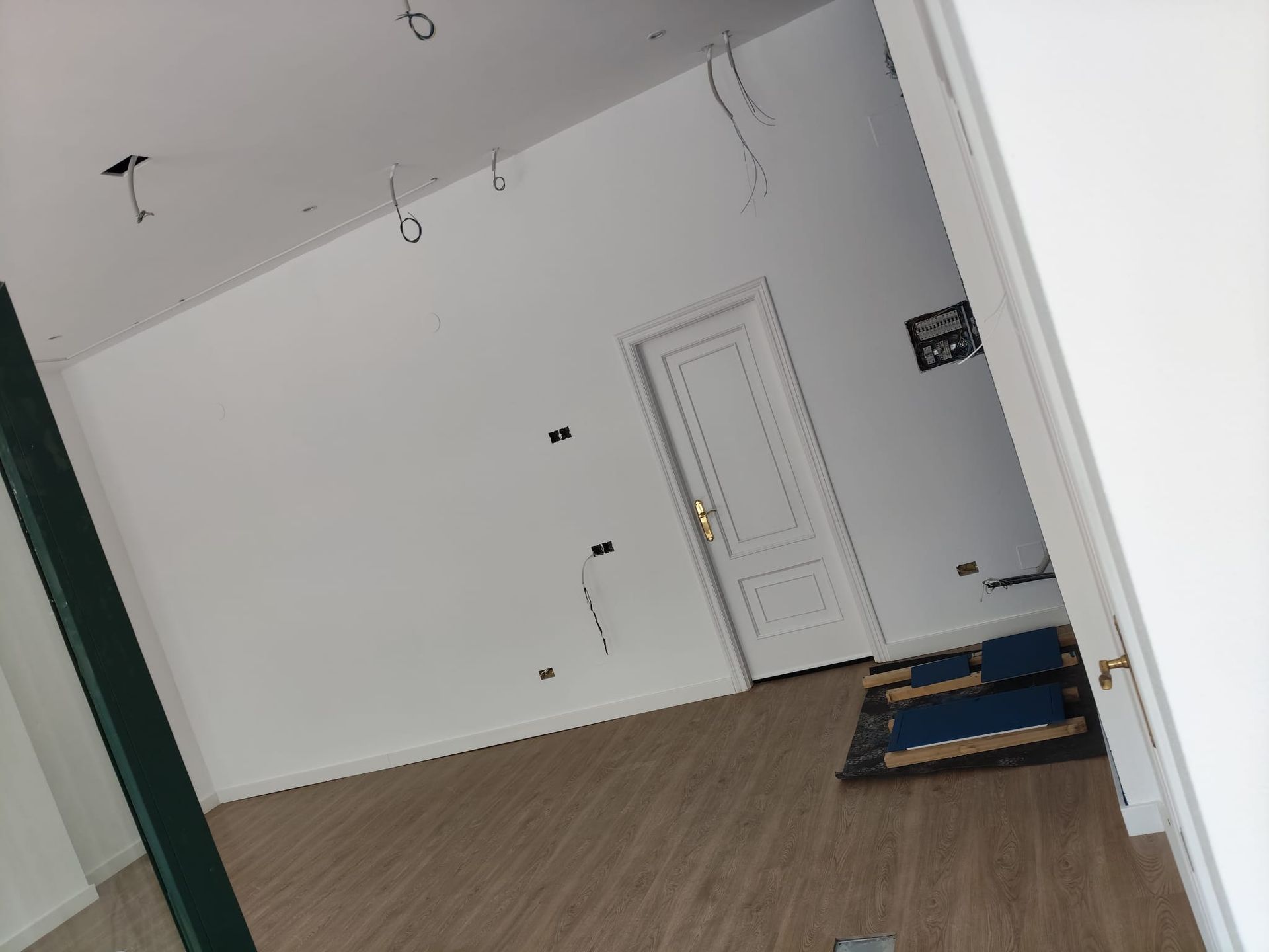 Habitación blanca vacía con suelo de madera, luces en el techo, una puerta cerrada y una alfombra azul junto a la pared.
