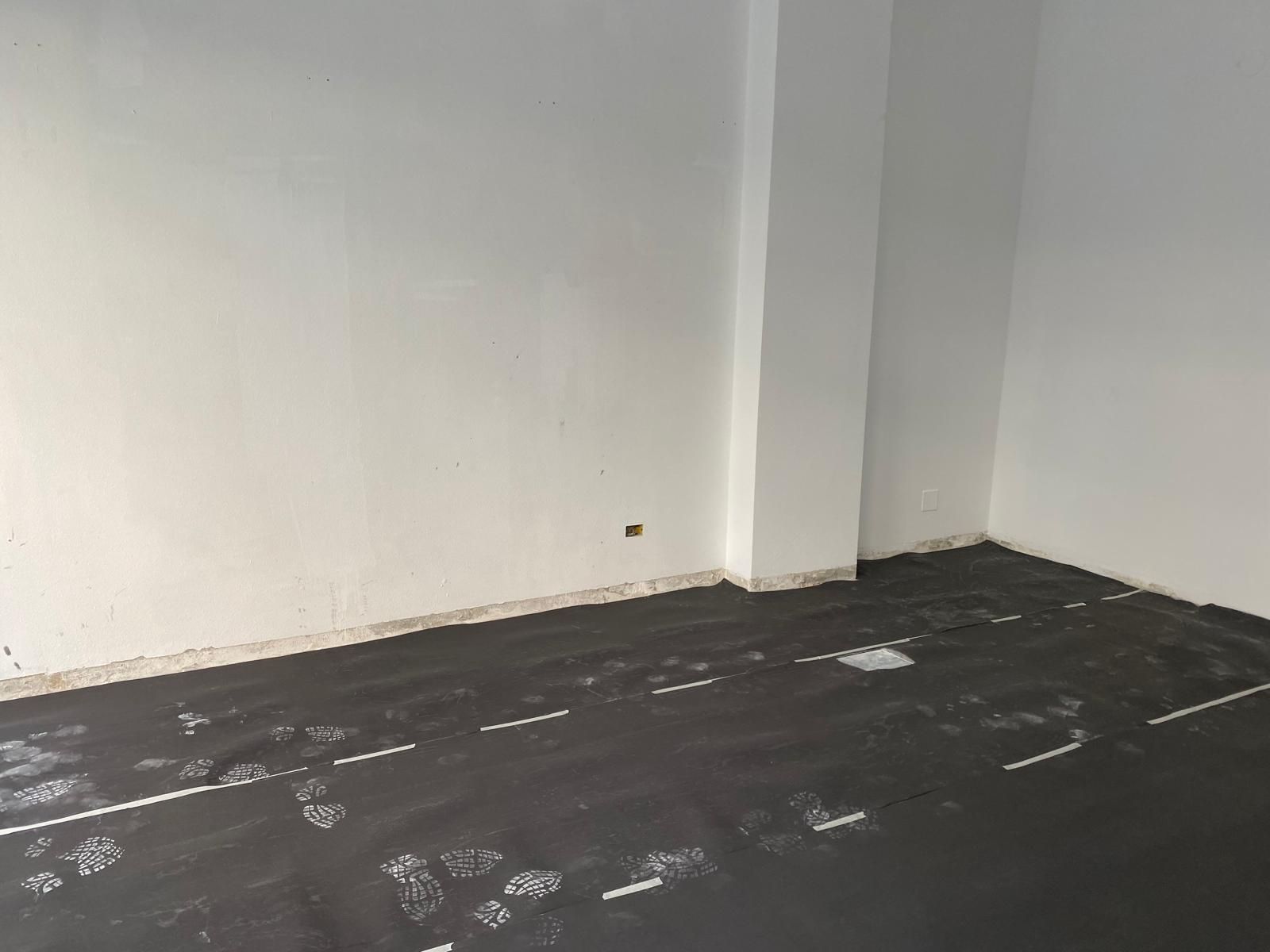 Rincón vacío de una habitación con paredes blancas y suelo oscuro marcado con líneas de tiza blanca o pintura.
