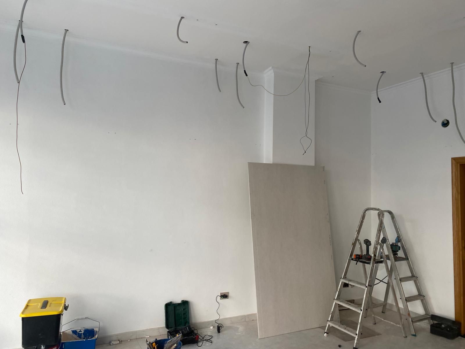 Habitación vacía en obras, con cables colgando, una escalera y una tabla apoyada contra la pared.