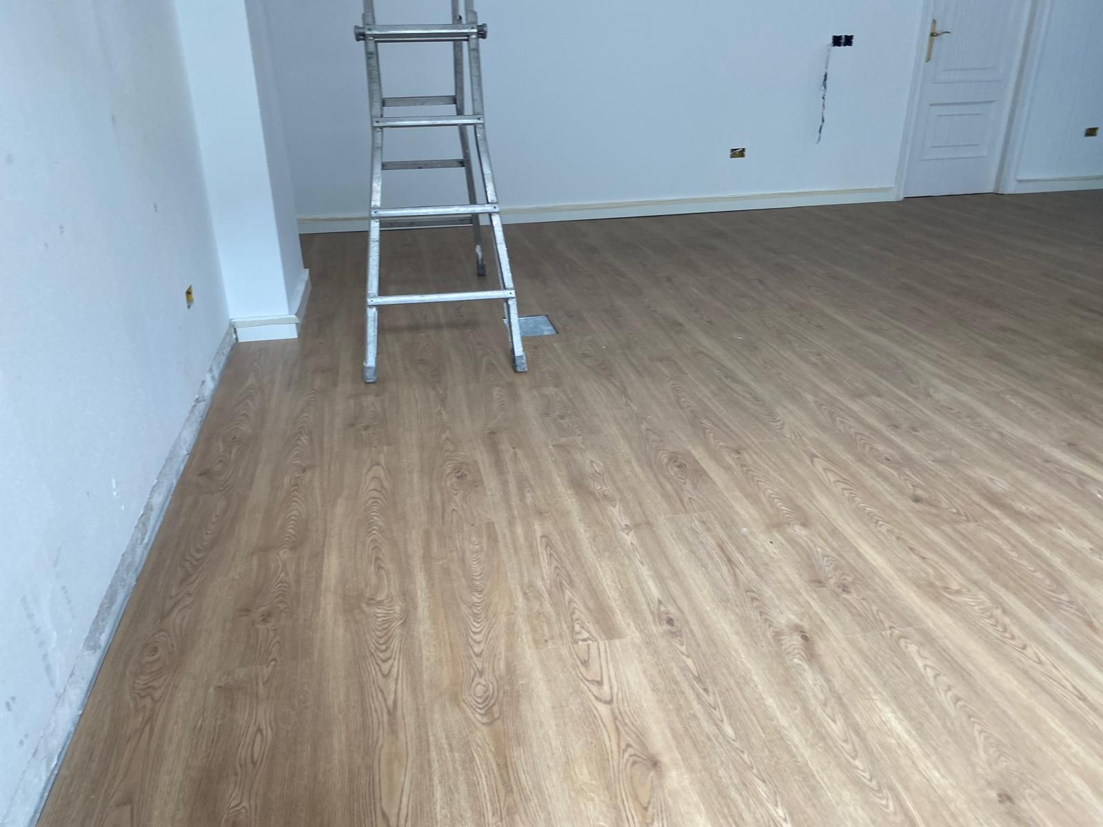 Habitación vacía con suelo de madera clara y una escalera cerca de la pared del fondo.