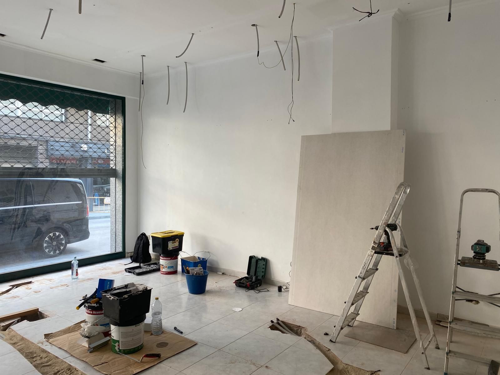 Cuarto de renovación con paredes blancas, cableado de techo suspendido, herramientas, escalera y materiales de pintura cerca de una ventana grande.