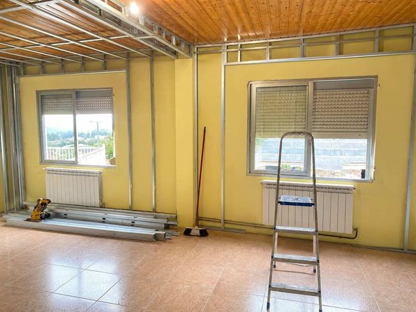 Habitación vacía en obras con paredes amarillas, techo a la vista, ventanas, escalera y suelo de material nuevo.