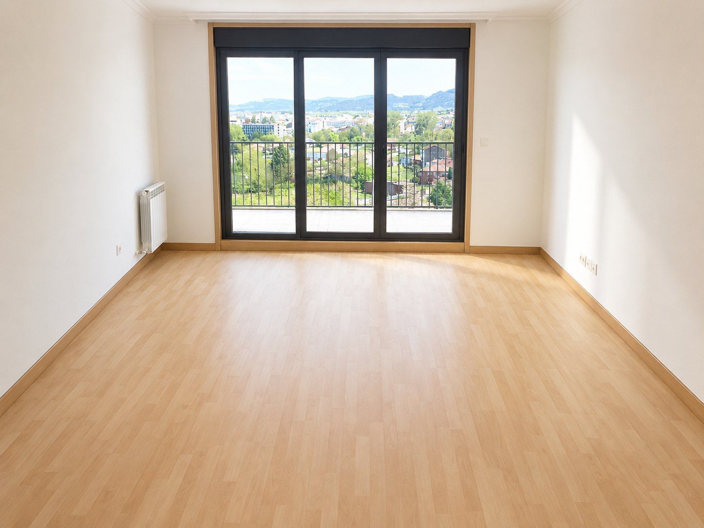 Habitación vacía con suelo de madera clara, paredes blancas y grandes puertas de cristal que dan a un balcón con vistas.