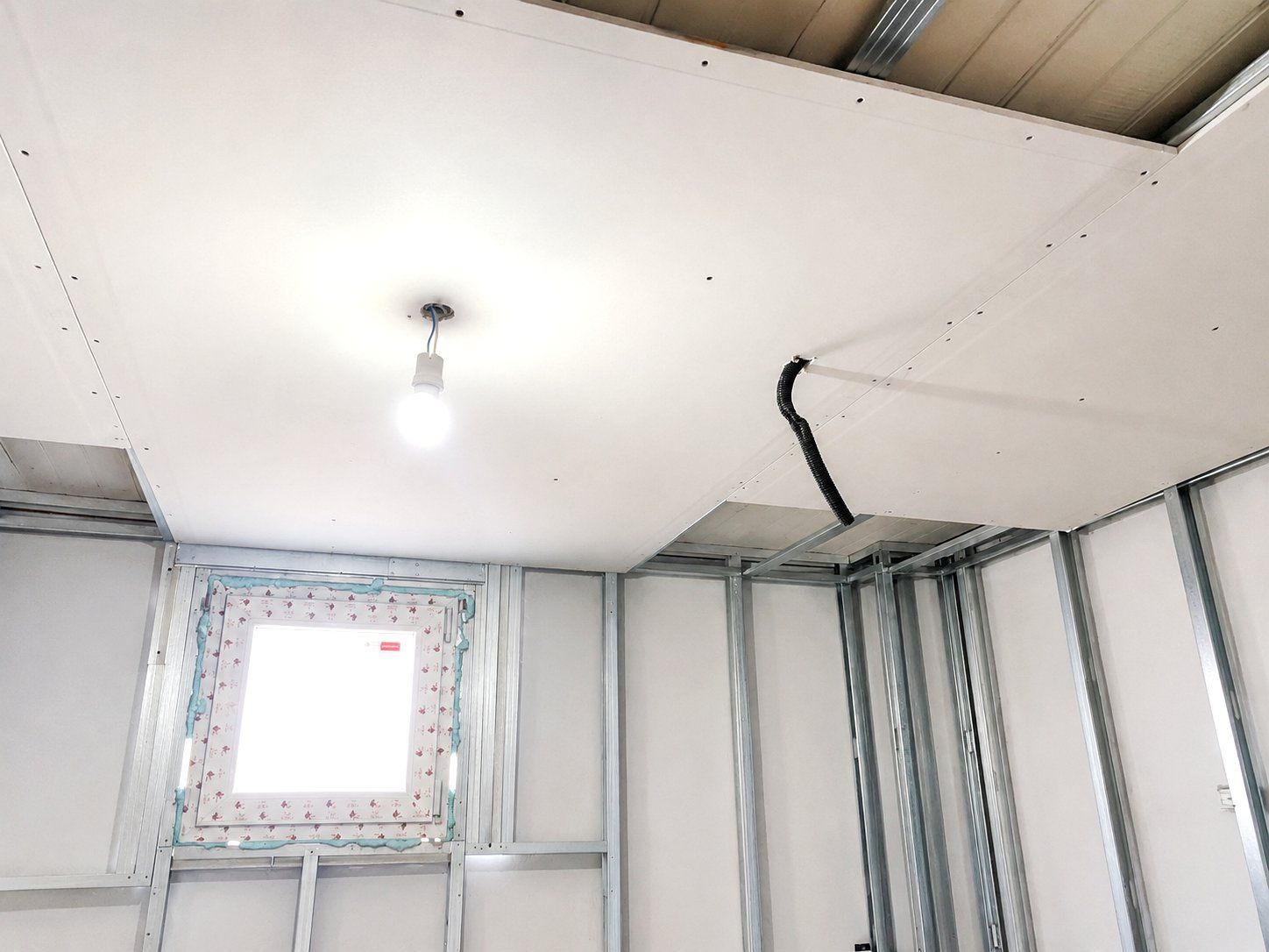 Habitación parcialmente terminada con paredes blancas, montantes metálicos, una bombilla al descubierto.