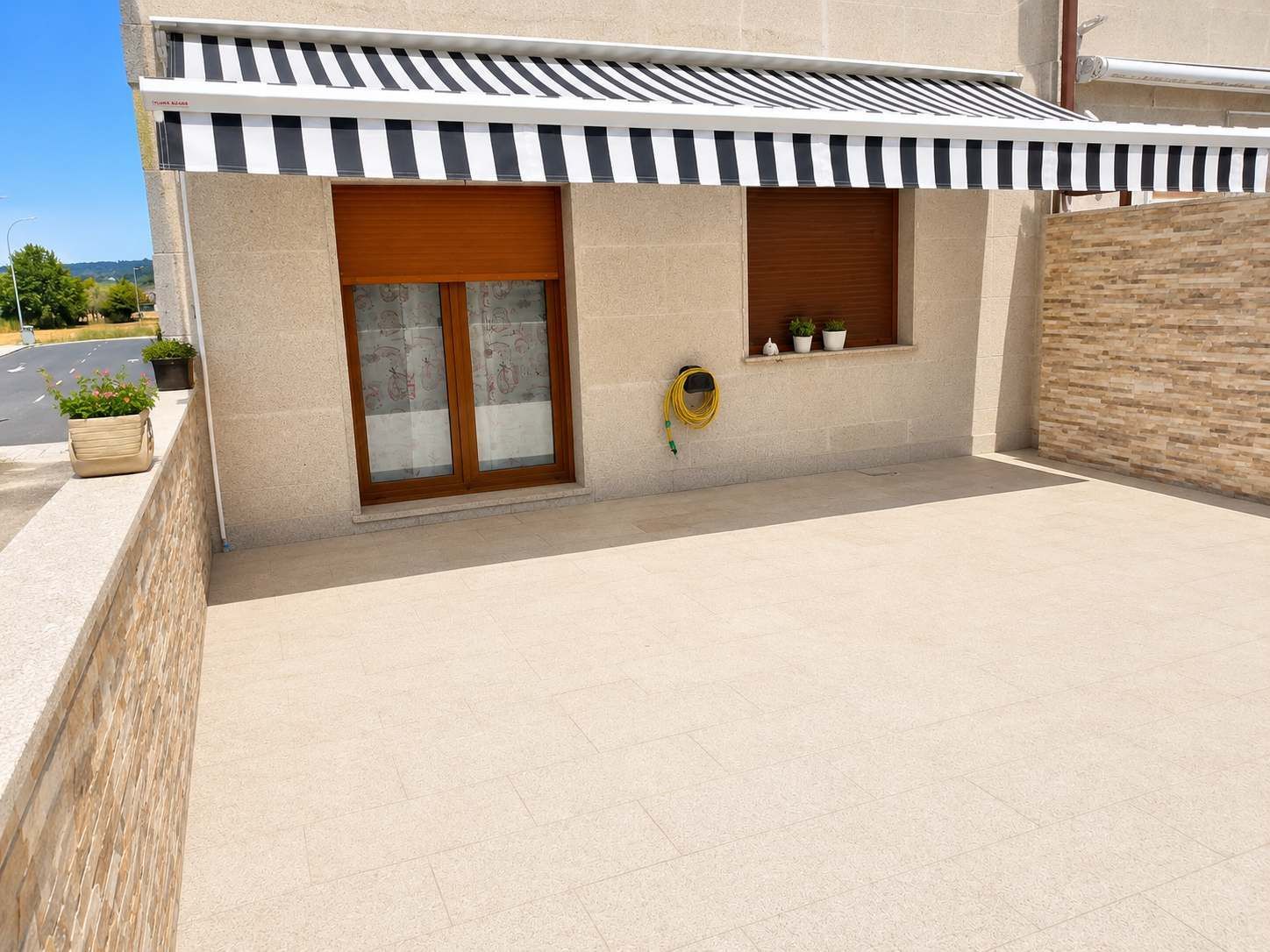 Pequeño patio embaldosado con toldo a rayas, puerta corredera de cristal, ventana y muro de piedra.