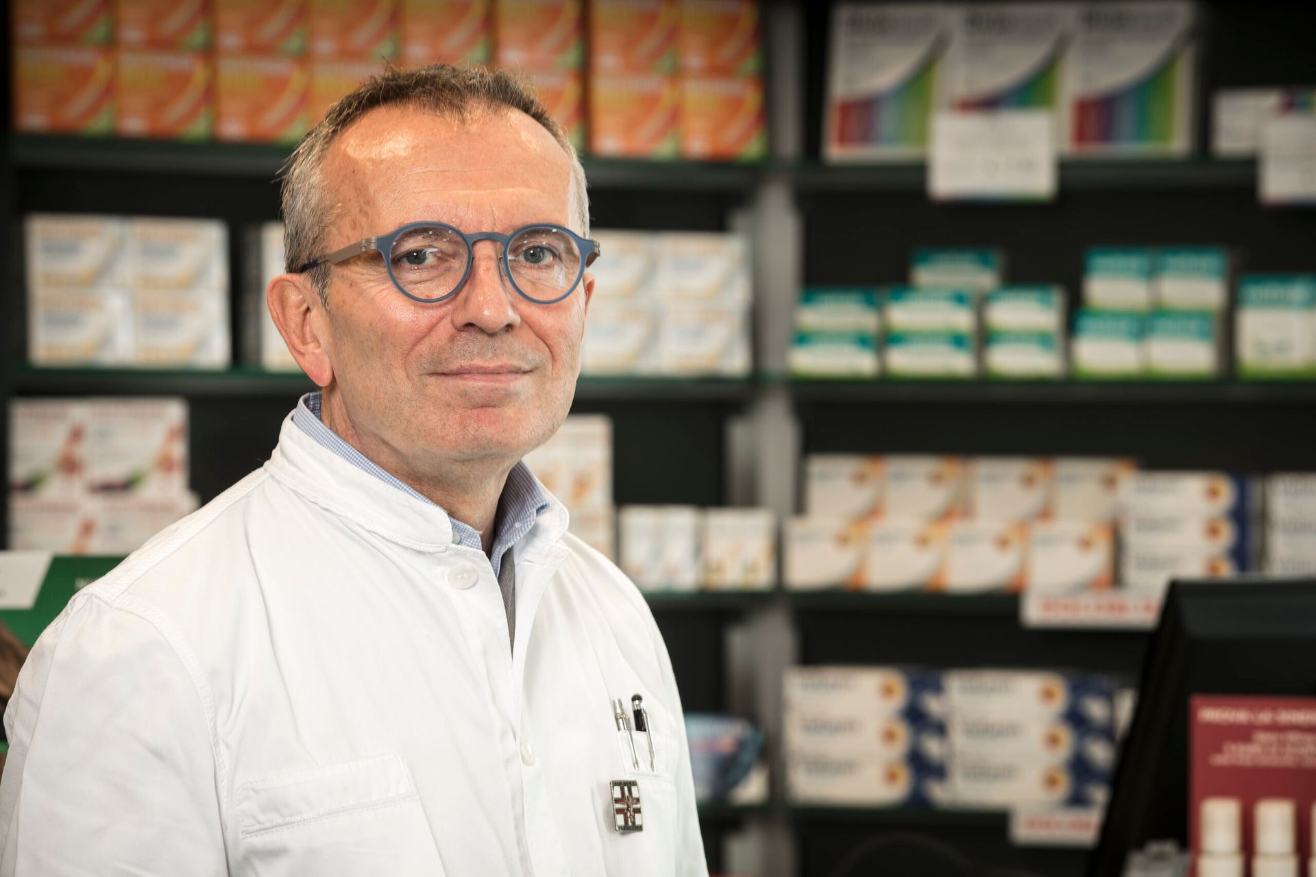 Un farmacista in camice bianco e occhiali sorride in una farmacia, sullo sfondo scaffali di medicinali.