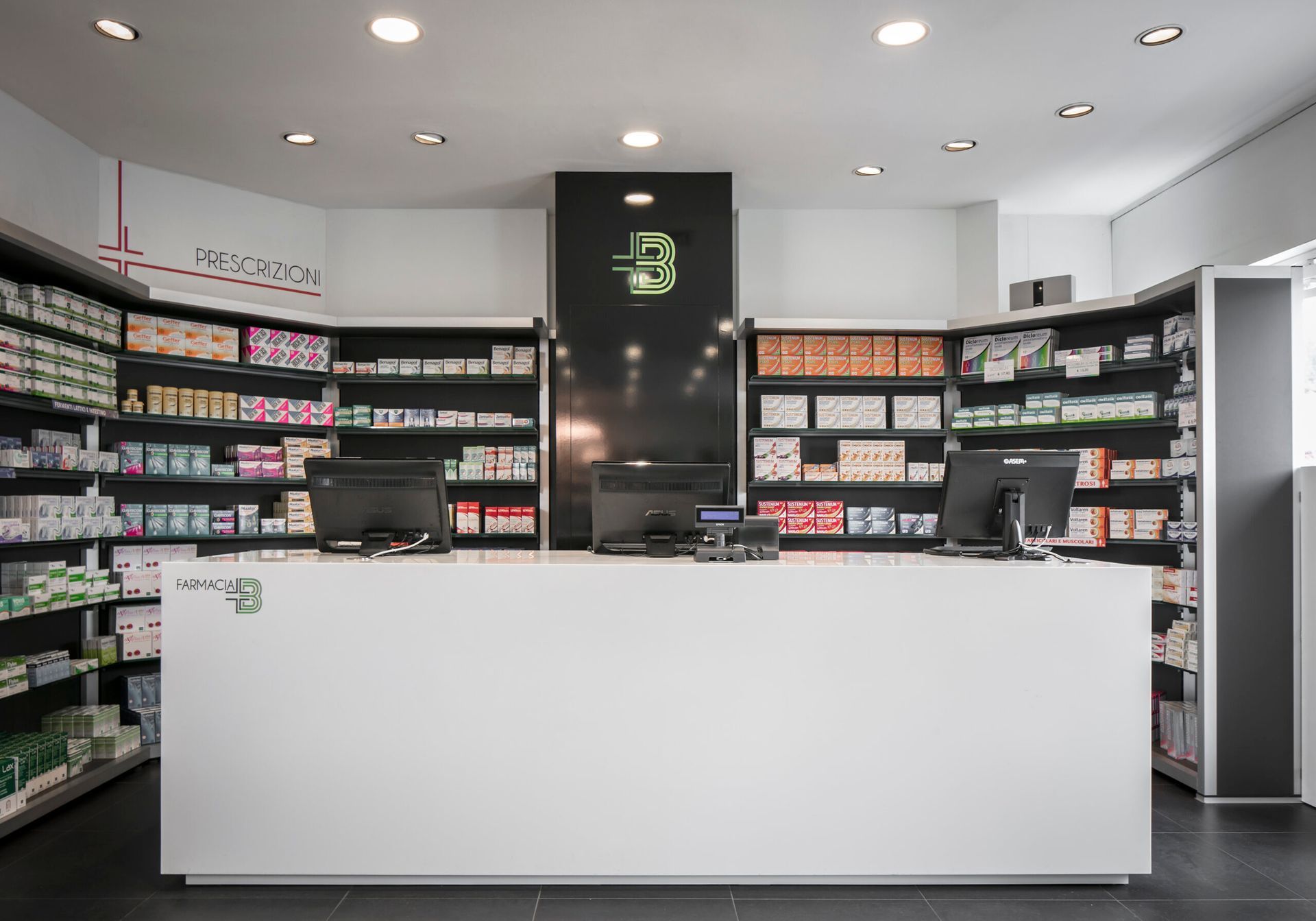 Interno di una farmacia moderna con bancone bianco, scaffali con medicinali e monitor per computer.