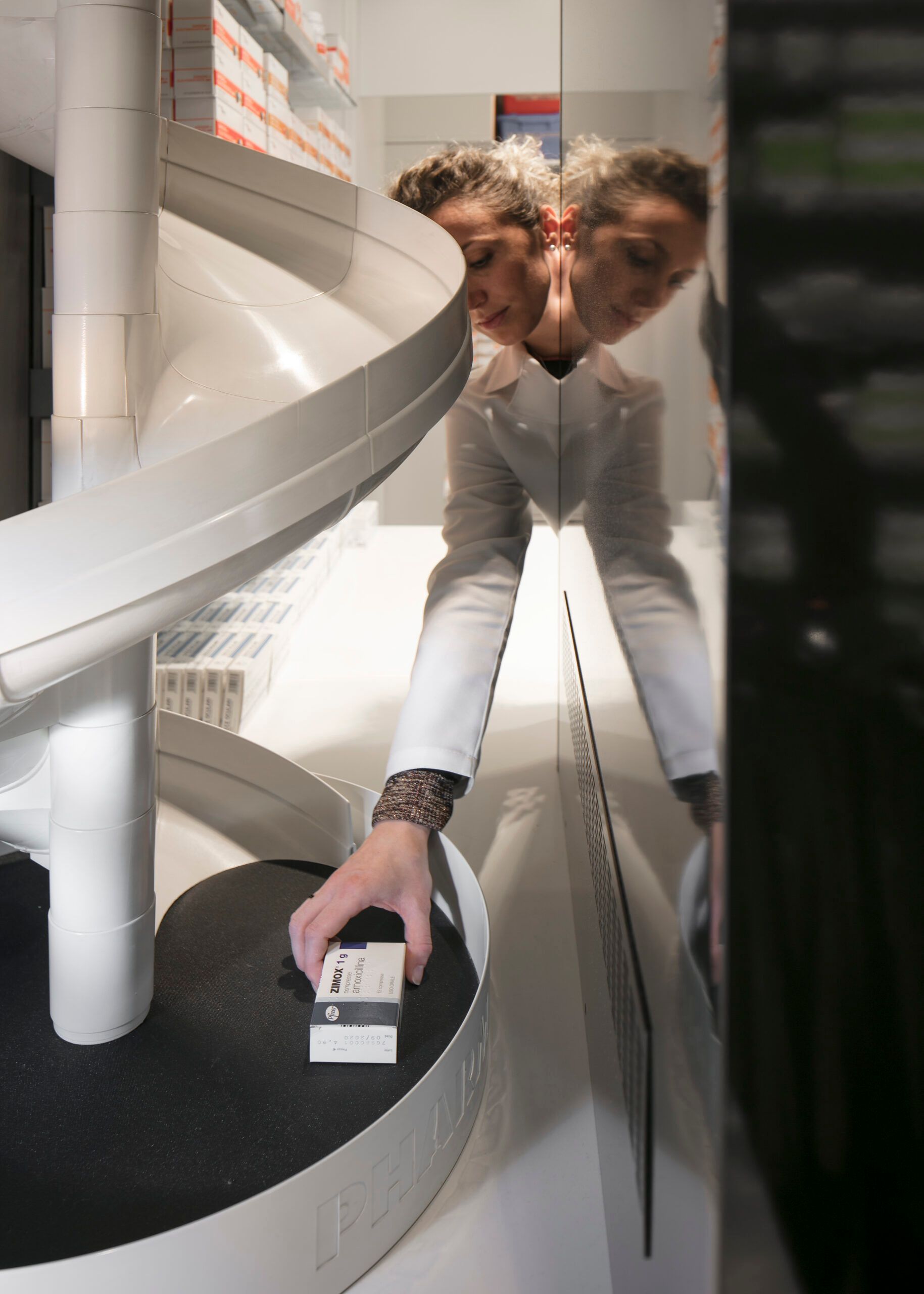 Donna in camice da laboratorio che infila la mano nel sistema di stoccaggio automatizzato di una farmacia.