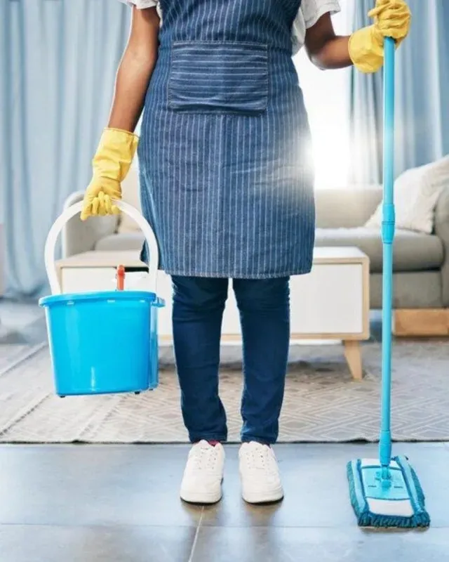 Persona que usa guantes y delantal sosteniendo suministros de limpieza en una sala de estar.