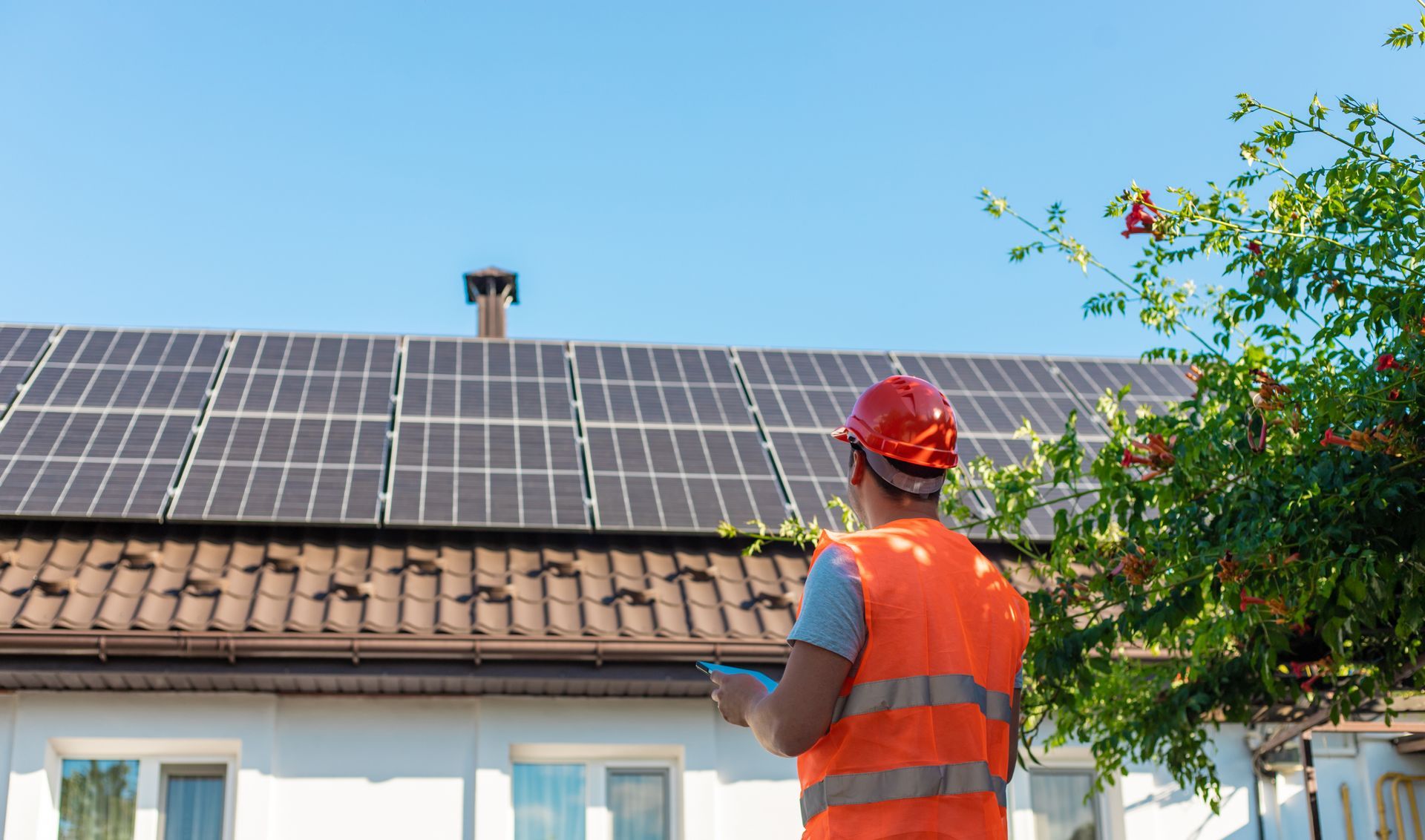 Arbeiter in Sicherheitsweste inspiziert Solarmodule auf einem Dach unter blauem Himmel.