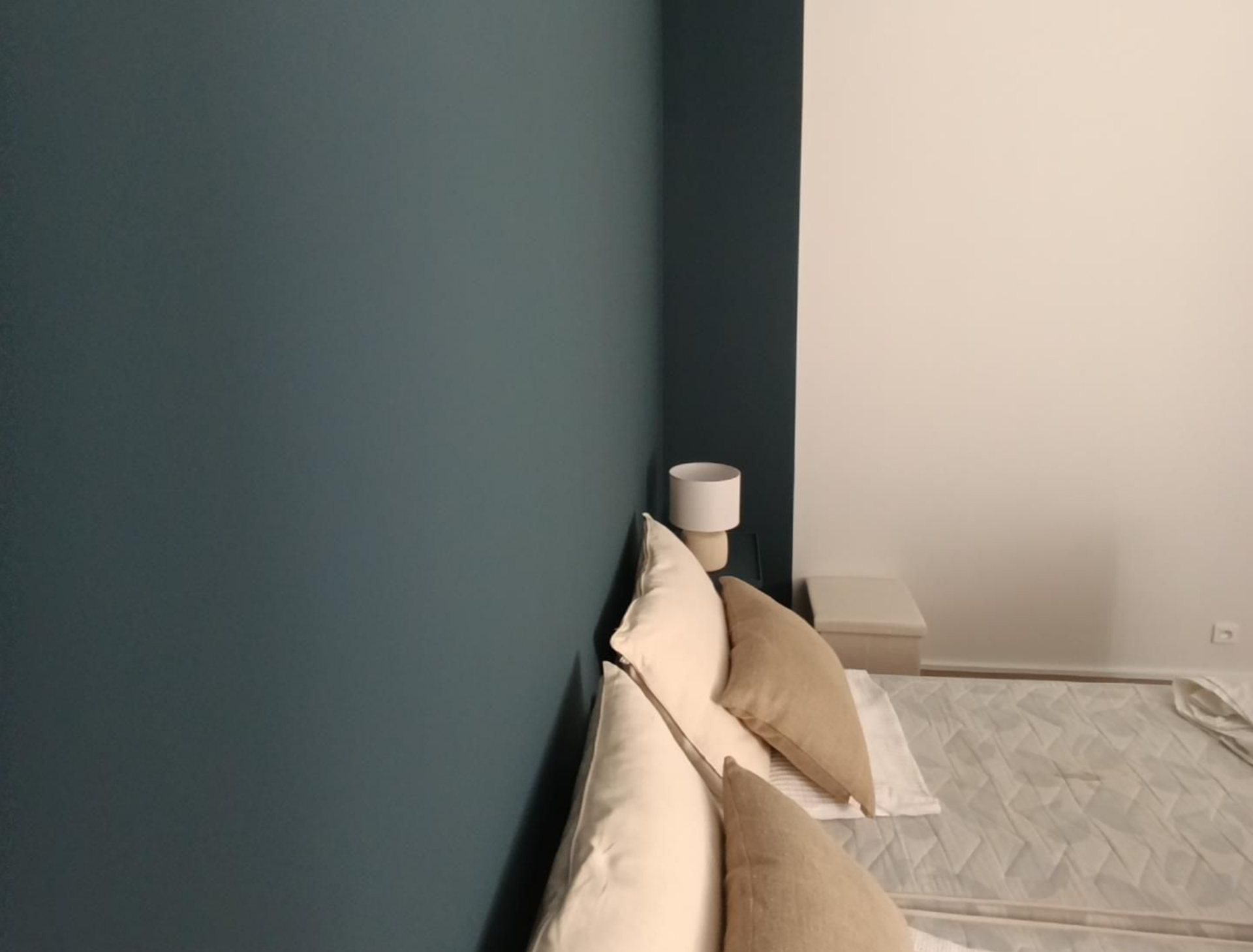 Coin de chambre bleu sarcelle foncé et blanc, lit avec oreillers beiges, petite table de chevet avec lampe et tapis à motifs.