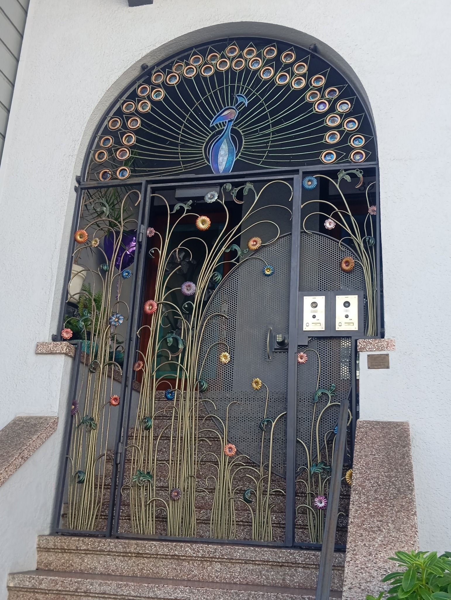 Portail en fer noir orné avec motif de paon, accents floraux et végétaux colorés.