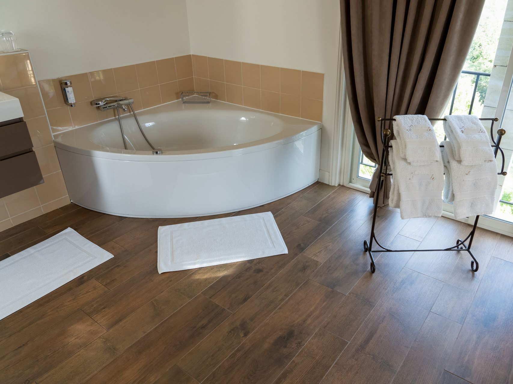 Salle de bains avec baignoire d'angle, serviettes sur un support et tapis blancs sur un plancher en bois foncé.
