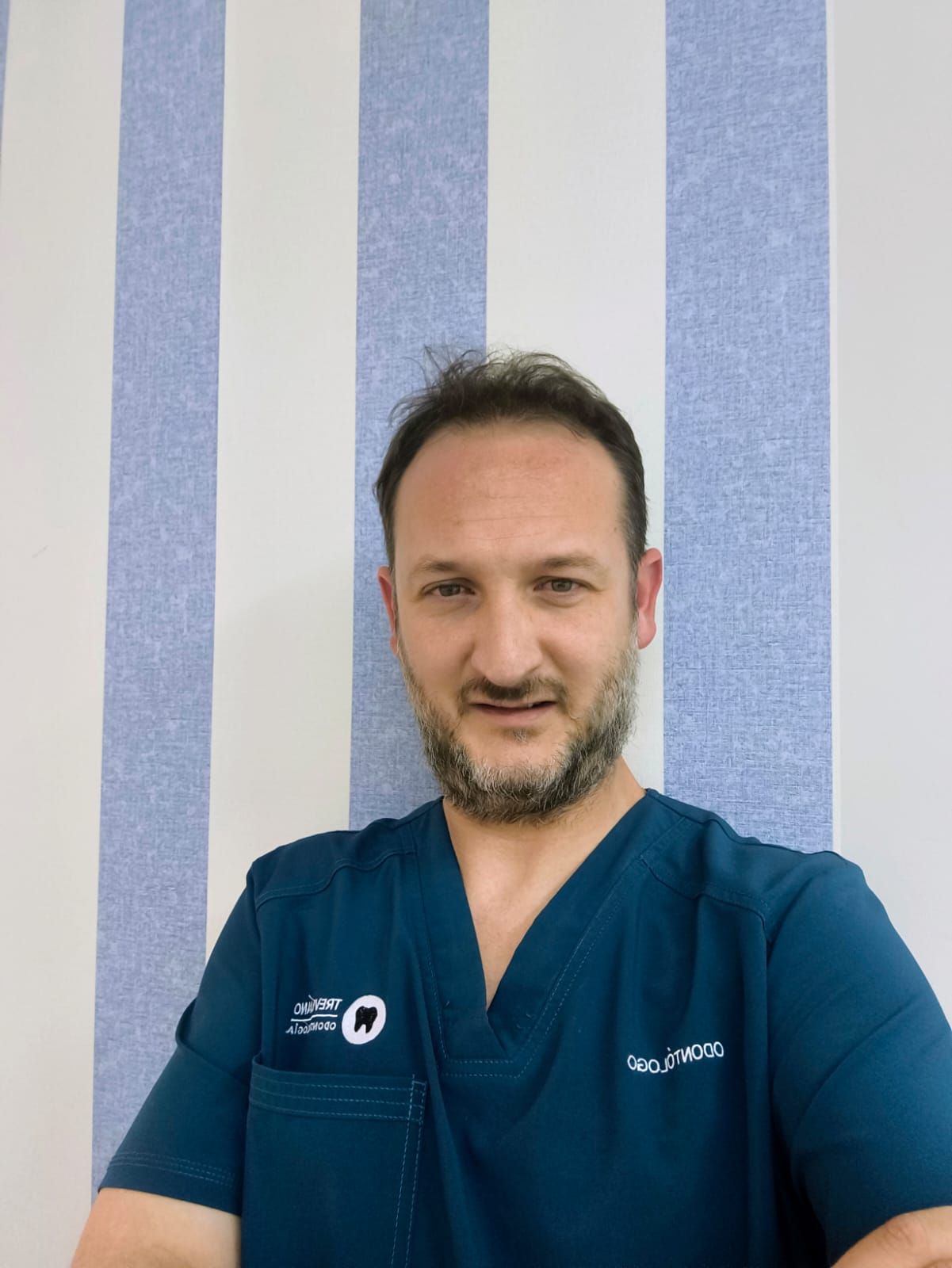 Hombre con bata médica color turquesa de pie contra una pared blanca lisa, mirando a la cámara con expresión neutra.