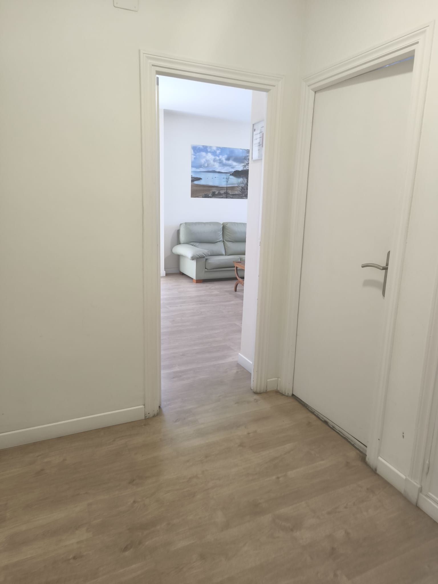 Pasillo blanco con dos puertas; a través de la puerta central se ve una luminosa sala de estar con un sofá gris.