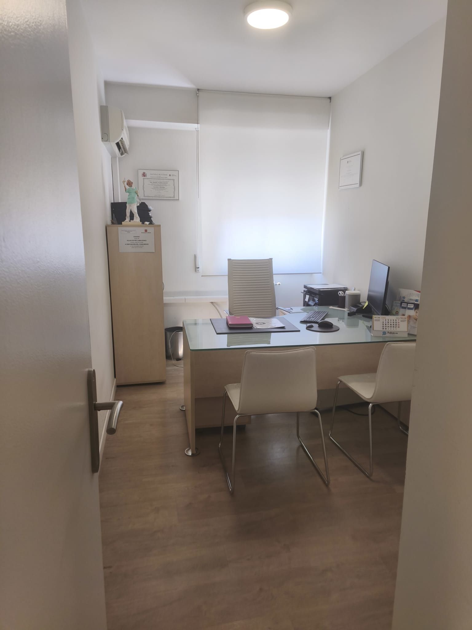 Pequeña oficina con escritorio, sillas, equipo informático y una ventana luminosa.