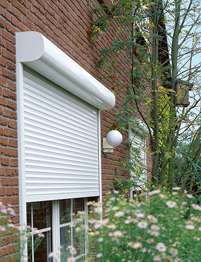 Ein weißer Rollladen ist über einem Fenster an einer roten Backsteinhauswand angebracht; im Vordergrund sind Grünpflanzen und weiße Blumen zu sehen.
