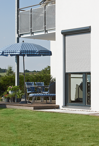 Eine weiße Außenwand mit einem teilweise geschlossenen Fensterladen, daneben eine Terrasse mit einem blau gemusterten Sonnenschirm und Stühlen.