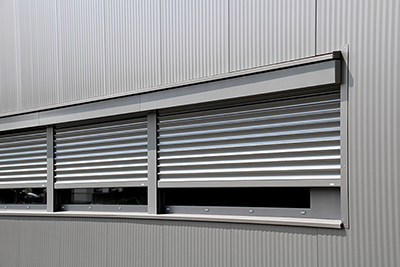 Eine moderne, metallische Gebäudefassade mit einer Reihe grauer, horizontaler Jalousien, die teilweise über die Fenster heruntergelassen sind.