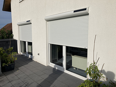 Zwei helle Fenster an der Hausfassade mit weißen Rollläden und integrierten Solarmodulen.