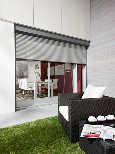 Vor einer Glasschiebetür, die teilweise von einem Rollladen verdeckt wird, befindet sich eine Terrasse mit einem dunklen Korbstuhl und einem Beistelltisch.