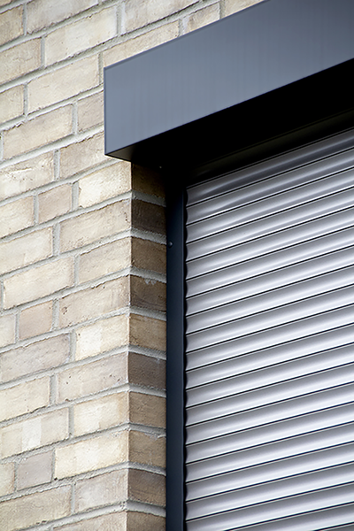 Nahaufnahme eines silbernen Rollladens, der in eine hellbraune Ziegelwand eingelassen ist, mit einem dunkelgrauen Kopfkasten und einer seitlichen Führungsschiene.