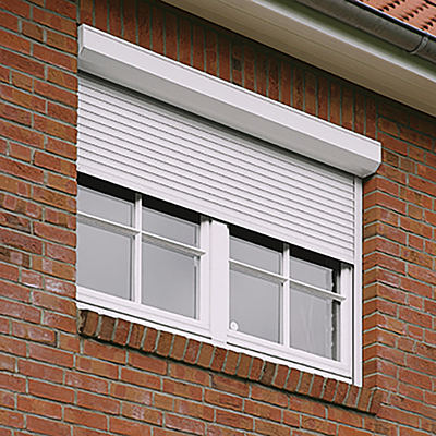 Ein weißer, außenliegender Rollladen ist über einem Fenster an einer roten Backsteinhauswand angebracht.