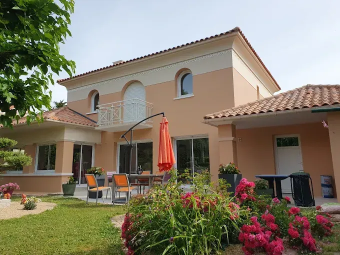 Une maison de deux étages couleur pêche avec un toit de tuiles rouges, du mobilier de patio et un jardin avec des fleurs rouges au premier plan.