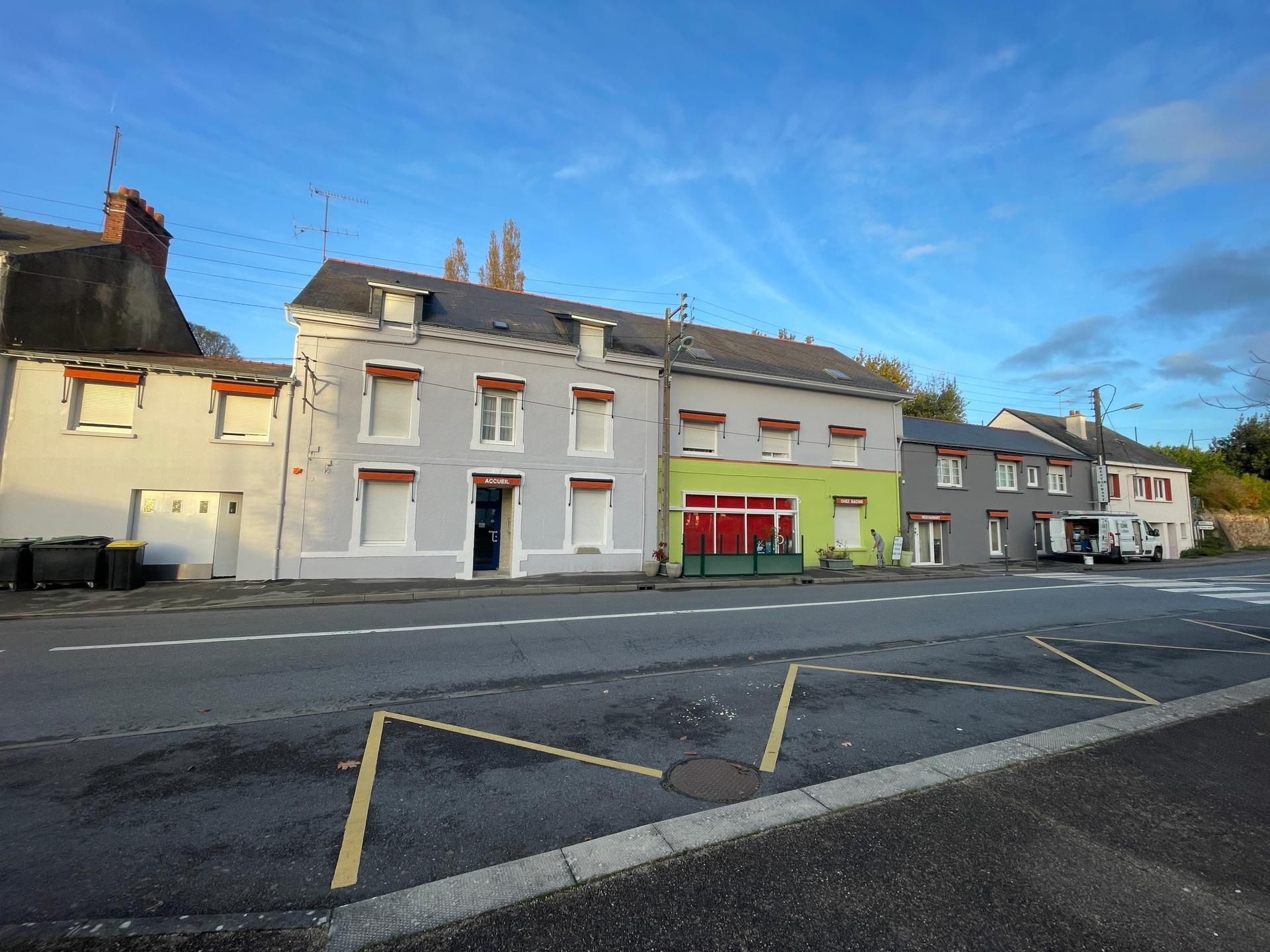 Une rangée de bâtiments le long d'une rue, dont une structure gris clair et une autre avec une façade vert vif sous un ciel bleu.