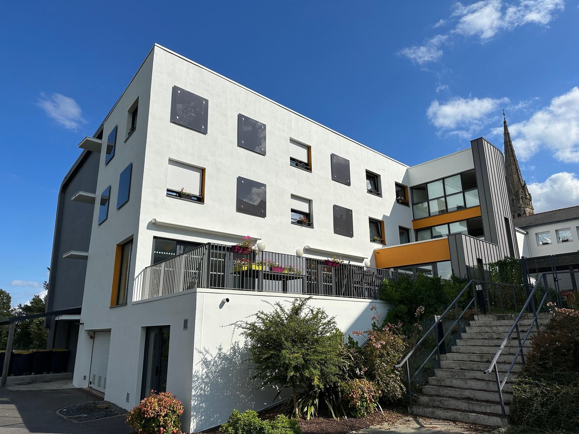 Un immeuble moderne blanc à plusieurs étages, avec de grandes fenêtres et un patio, se détachant sur un ciel bleu éclatant.