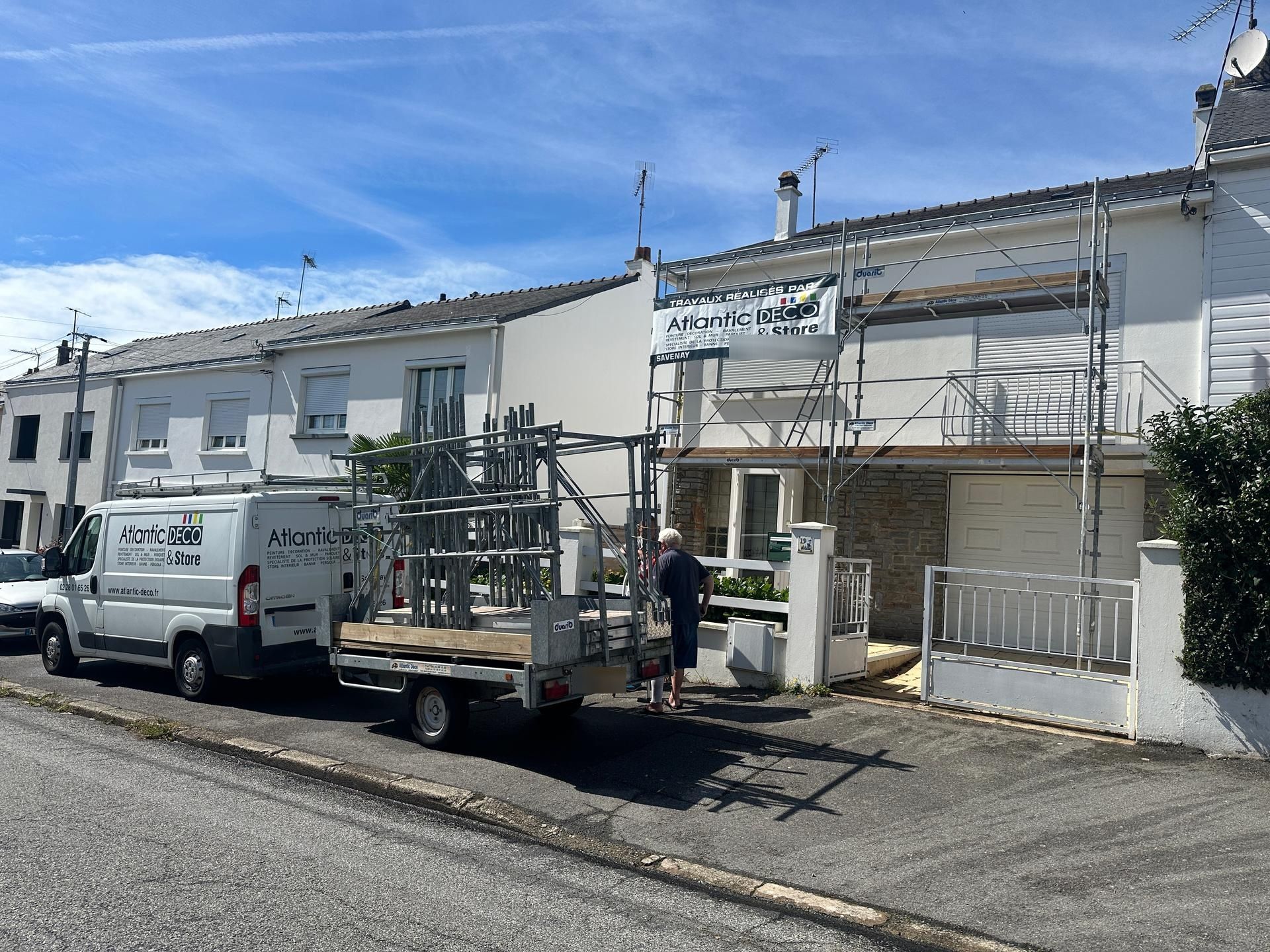 Une camionnette blanche avec une remorque transportant des échafaudages est garée devant une maison en rénovation.
