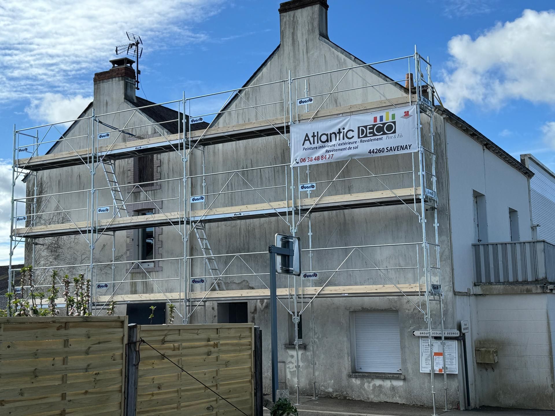 Un bâtiment de deux étages en rénovation, dont la façade grise et délabrée est recouverte d'échafaudages métalliques, sous un ciel bleu.