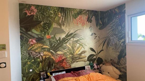 Un mur de chambre recouvert d'une fresque murale vibrante représentant une jungle luxuriante avec un dinosaure et un lit au premier plan.