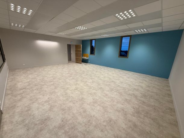 Un grand bureau vide, avec de la moquette gris clair, des murs blancs et un mur d'accent turquoise percé de deux hautes fenêtres rectangulaires.