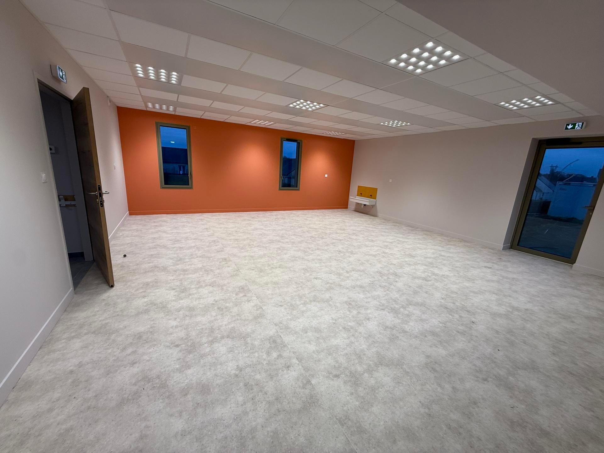 Une pièce vide, lumineuse et spacieuse, avec un sol gris clair, des murs blancs et un mur d'accent orange percé de deux fenêtres.
