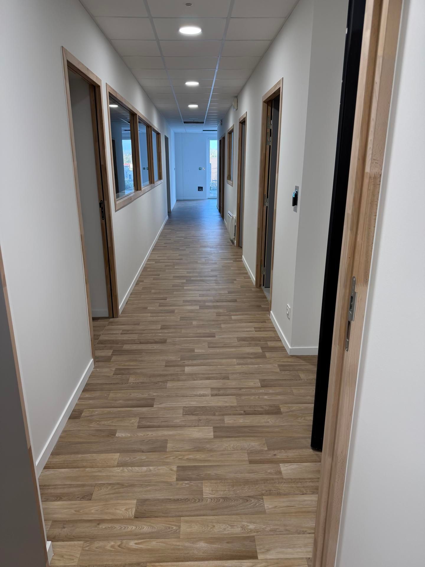 Un couloir de bureau lumineux avec un sol clair à motifs de bois, des murs blancs et de nombreuses portes à cadres en bois.