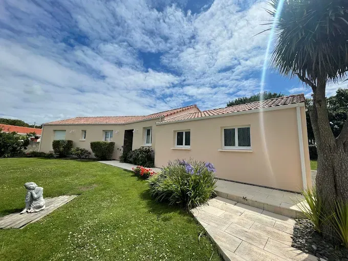 Une maison de plain-pied aux tons clairs, avec un toit de tuiles en terre cuite, entourée d'une pelouse et d'aménagements paysagers, par une journée ensoleillée.