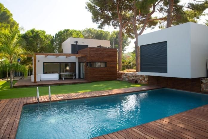 Casa moderna con piscina; exterior de madera y blanco, agua azul. Árboles al fondo, día soleado.