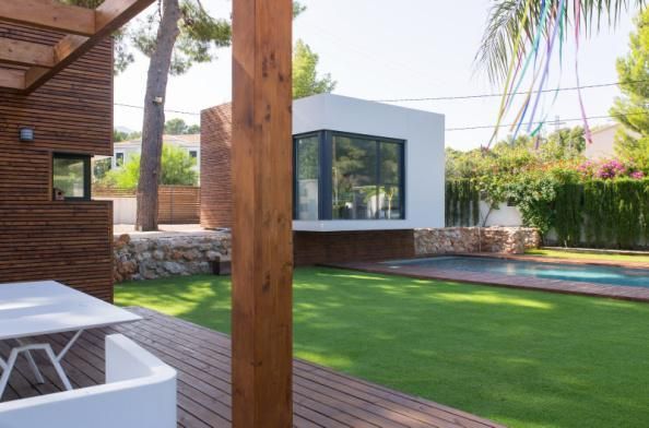 Casa moderna con revestimiento de madera, patio y césped, que cuenta con una estructura de cubo blanco independiente y una piscina.