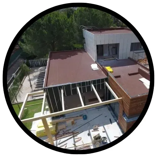 Sitio en construcción con techo parcialmente construido de color marrón y blanco, rodeado de árboles.