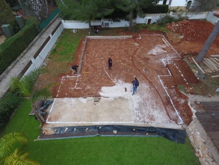 Obreros construyendo cimientos de hormigón al aire libre, parcialmente terminados. Suelo marrón rojizo, marcas blancas y césped verde.