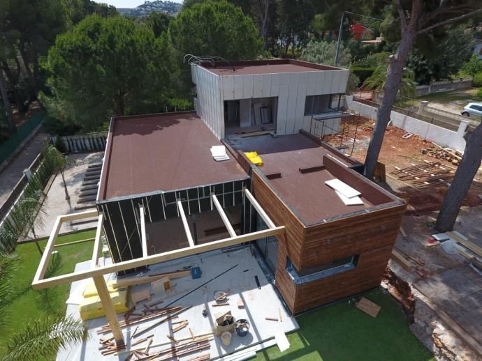 Vista exterior de una casa moderna en construcción con revestimiento marrón y beige, techo plano, rodeada de árboles.
