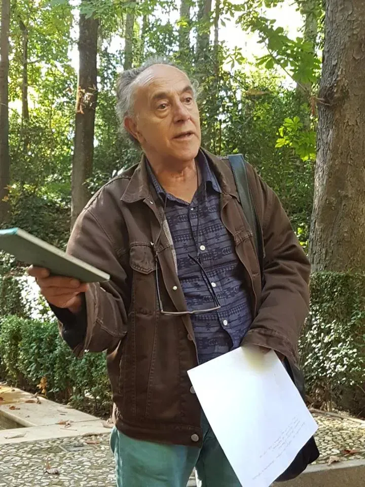 Persona de pie al aire libre en un sendero arbolado, sosteniendo papeles y una carpeta, mirando hacia arriba.