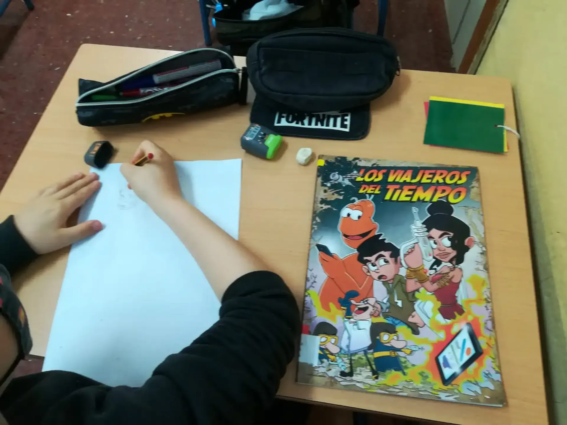Manos dibujando sobre papel blanco en un escritorio junto a un cómic colorido y estuches para lápices.
